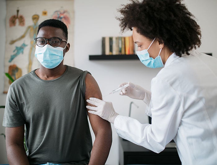 Stock photo of a black female health worker, wearing mask and scrubs, standing over a black male patient and adminsters a shot in arm