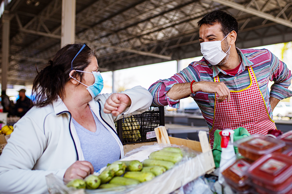 Prevention and Wellness. Two people wearing masks in a farmers market exchange greetings by bumping elbows.