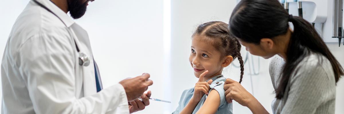Stock photo of a young girl, sitting with her mother, smiling up at doctor holding a syringe