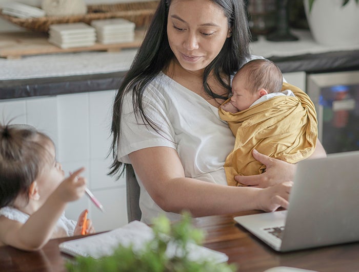 stock photo of mother seated at a table with a laptop and notepad. Woman is holding an infant and engaging with a toddler