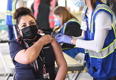 Masked woman holds up sleeve while health worker applies bandage to her shoulder