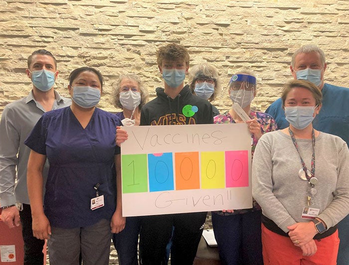 Health workers wearing masks stand around posterboard that reads '10000 vaccines given!!''