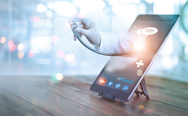 3 Takeaways from the Rise of Virtual-First Health Plans. A clinicians hand holding the bell of a stethoscope reaches through the screen of a tablet displaying a telehealth app.