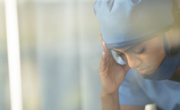 How to Help Clinicians Cope with the Pandemic’s Impact. A physician in scrubs has a hand on her head and is looking down.