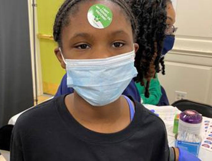 Little girl with braids wears a black teeshirt and a sticker on her forehead at a vaccination event.