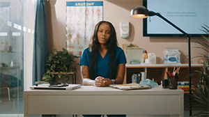 Black female health worker in scrubs sits behing a desk staring seriously into camera