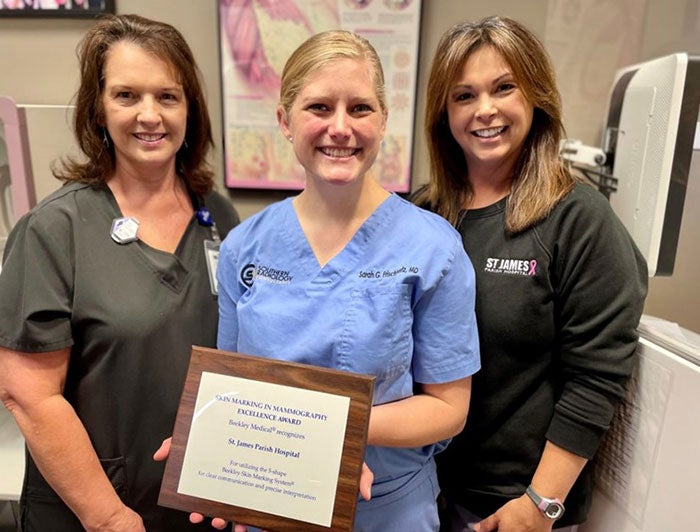 Three women posing for a picture, smiling, and wearing scrubs while the woman in the middle is holding a plaque award.