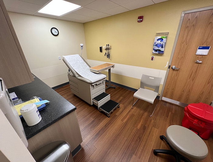 Patient room at a doctor's office with a reclining bed chair, rolling chair, counter with sink, hazard waste basket, and door for entry/exit.
