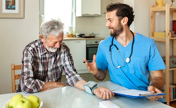 4 Ways a Signify Health Deal Could Help CVS Health. A home health clinician with a clipboard to record health data meets with an elderly man who is wearing a monitor on his left wrist at his kitchen table..
