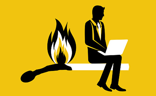 Executive Burnout Is Real — and It Can Be Reduced. An executive in a business suit sits on the unburned end of a matchstick working on his computer as the matchstick burns down towards him.