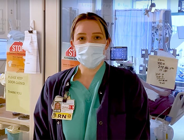Female RN in scrubs and badge stands in front of patient room in hospital
