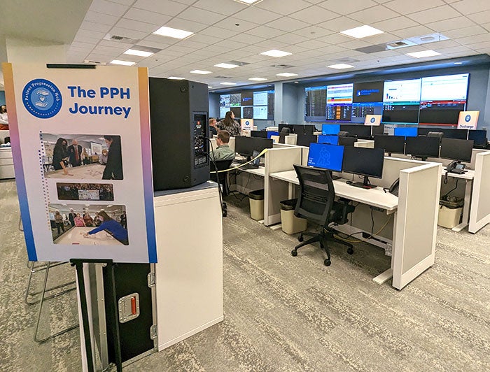 Photo of cubicles facing a wall of screens with a sign standing on the left side of image reading 'The PPH journey'