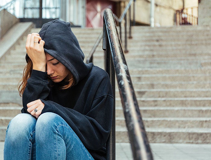 Stock photo of an adolescent girl wearing hoodie and looking upset, sitting outside on rundown building steps