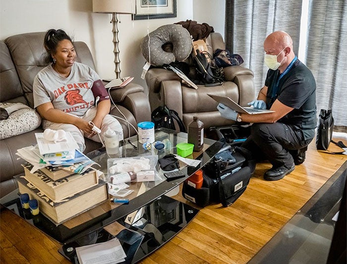 A bald-shaven paramedic kneels in a patient's living room, checking her blood pressure
