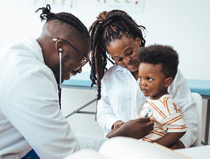 Stock photo of a black physician smiling and examining a black toddler sitting on his mother's lap