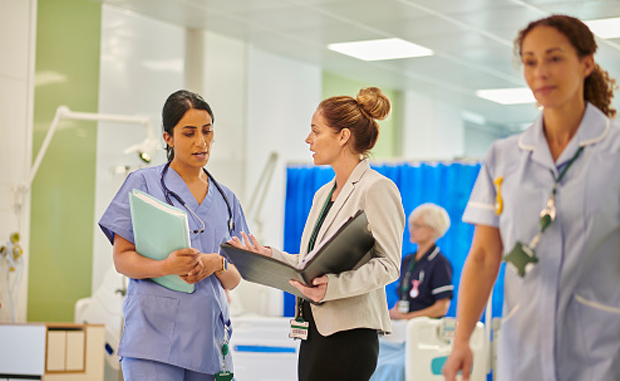 6 Ways to Build a Rewards Strategy to Retain Nurse Managers. A nurse leader speaks with a hospital executive about nurse manager retention policies on a hospital floor.