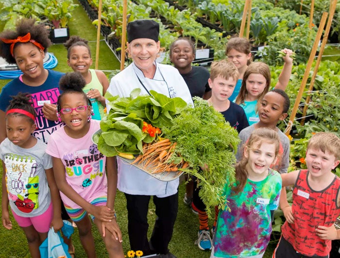 A chef stands in a garden holding produce, surrounded by smiling children