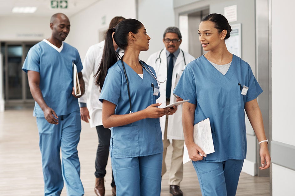 Workforce. Health workers, some wearing scrubs and some white coats, talk to each other as they stroll through a hospital hallway