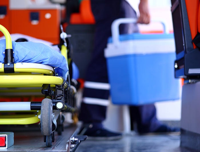 Stock photo of EMT carrying an organ cooler in the background, gurney in foreground