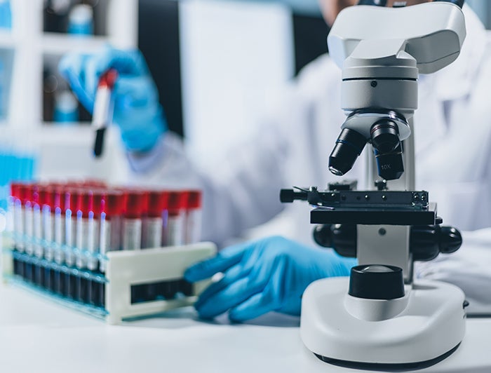 West Virginia University Medicine Children’s. Stock image of scientist in a lab behind a microscope, holding test tube. 