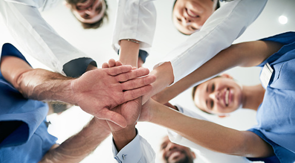 4 Ways Updating Workforce Models Can Deliver Big Results. A group of clinicians gathered in a circle put their hands on top of each other's hands as part of a team celebration.