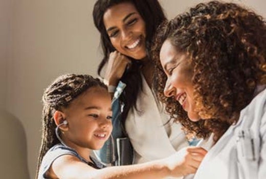 A pediatric patient listens to a physician's heartbeat through a stethoscope while her mother watches with a smile on her face.