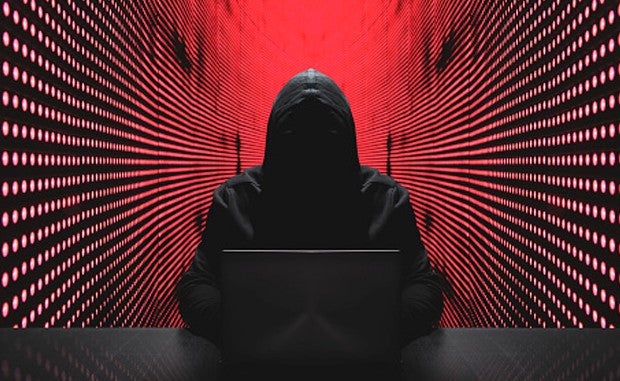 Third-Party, Cyber-Risk Skyrockets for Health Systems. A hacker in a black hoodie that leaves his face in shadow sits in a room lit by red lights and works on a laptop to break into a health system's computer network.