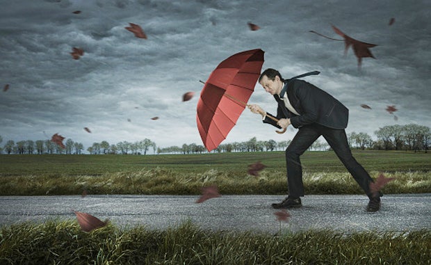 What’s Driving Headwinds for Insuretechs? A man in a business suit holding a red umbrella walks on a sidewalk into high winds during a storm.