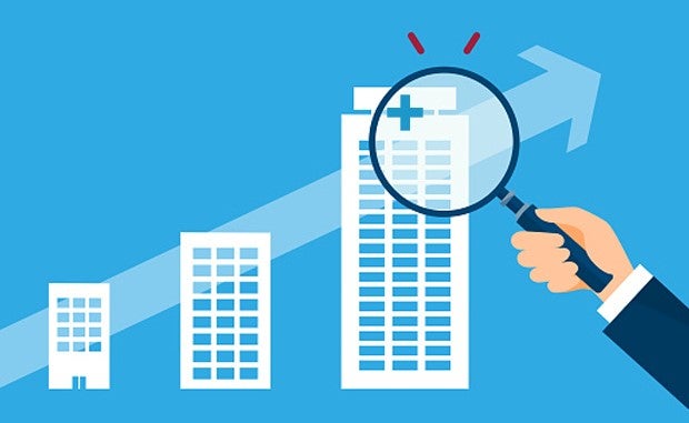 Why General Catalyst Is Doubling Down on Health Care Investments. Three buildings of increasing size with an upward pointing arrow overlaid on them. A business person's arm is shown holding up a magnifying glass to the top of the tallest building and blue hospital cross is visible.