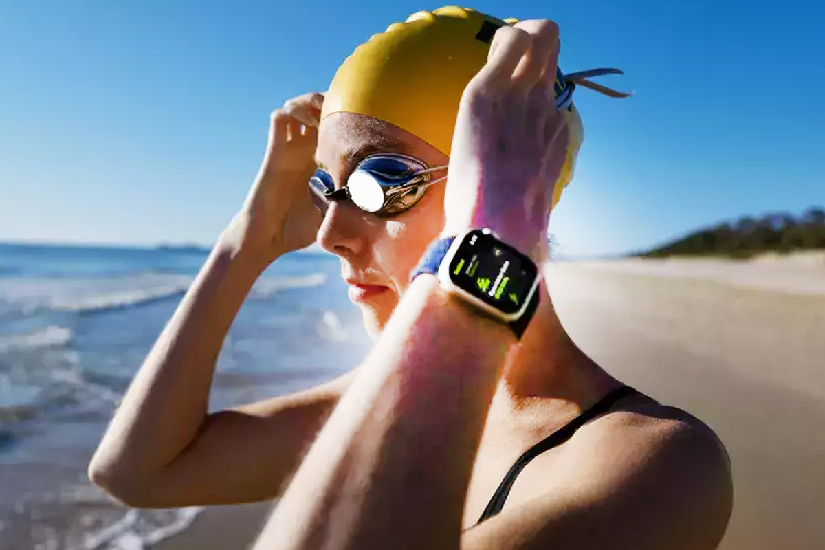 Will Employees Become Fitter and Share Wearable Device Data for $1,000 a Year? A woman on a beach wearing a swimsuit, swim cap, swim goggles, and a fitness tracker.