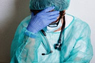 Many Workers Have Yet to Get Mental Health Care They Need Due to Pandemic. A clinician in a robe, a mask, glasses, and gloves holds her head in her hand.