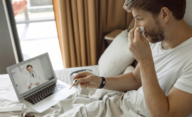 Consumer Attitudes Changing on Accessing Care Remotely. A man at home in bed with cold symptoms and holding an electronic thermometer talks with a clinician on his laptop computer via a telehealth application.