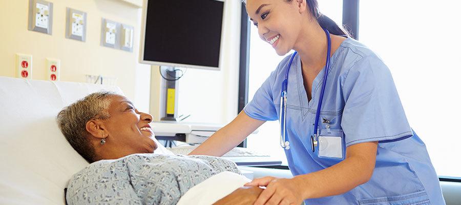 Partnering on Population Health. A Black female patient in a hospital bed smiles at a female clinician of color with a smile on her face who is examining the patient in a hospital room.