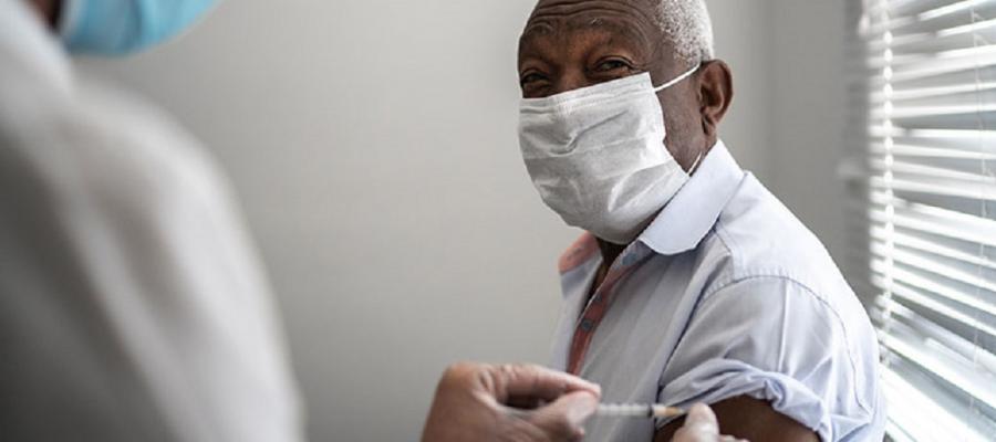 COVID-19 Vaccine. A elderly man of color wearing a mask receives a COVID-19 vaccine in his right arm from a masked, gowned, and gloved clinician. Vaccine images.