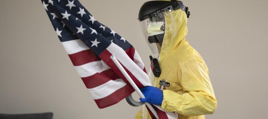Living Learning Network. A person in a hazmat suit wearing rubber gloves and a mask under his face shield holds an Unites States flag.