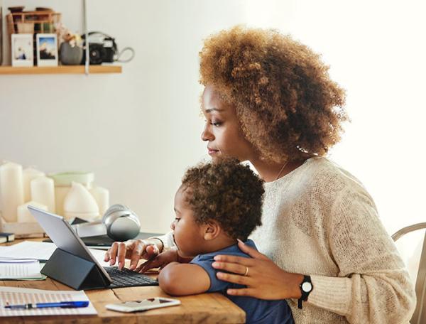 A mother sits at a dining table with a toddler on her lap, staring at a tablet computer screen