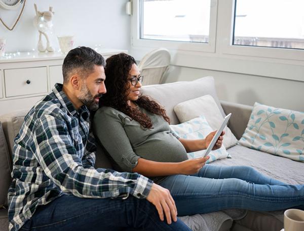 A pregnant couple sits in a room watching a roundtable on a tablet