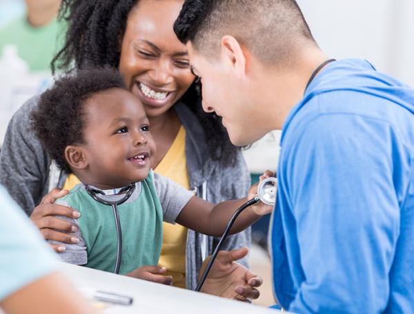 Stock photo shows toddler seated on mother's lap during an examination, wearing clinician's stethoscope