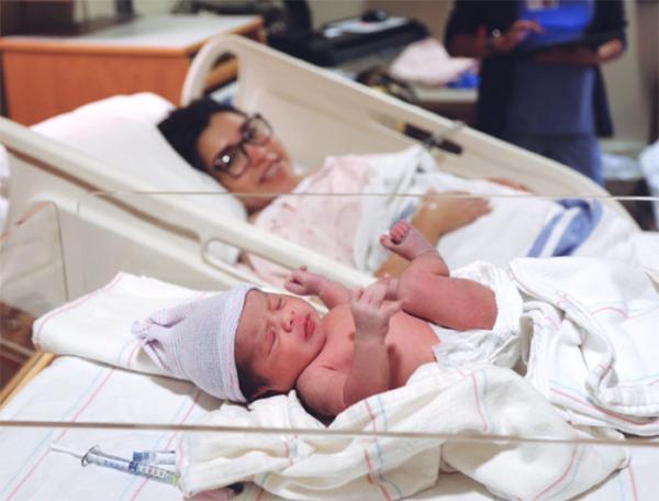 A newborn sleeps in a hospital bassinette next to a mother in a hospital bed