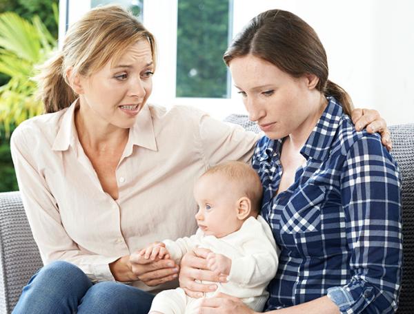 A woman sits on a sofa comforting a sad mother holding a baby