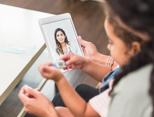 A mother sits with child on her lap having a virtual consult with a clinician shown on her tablet