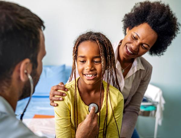 A preteen girl sits with her mom behind her as a clinician checks her heart her 