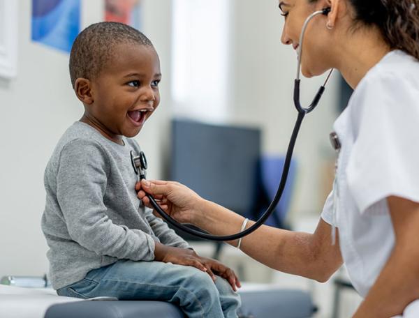 A little boy is seated on a table smiling at the female clinician examining him with a stethoscope