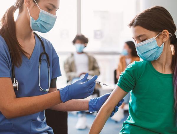 A tween girl wearing a surgical mask gets a shot from a female clinician in scrubs and surgical mask