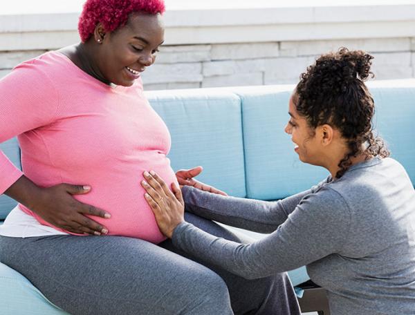 A pregnant Black mother-to-be sits as a Black nurse or doula touches her stomach