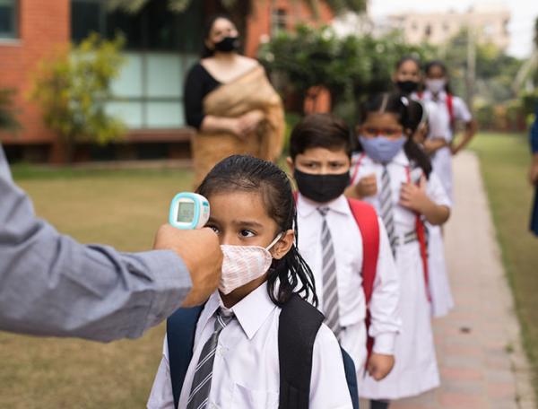 children in school uniforms and face masks line up outside school to have temperature checked