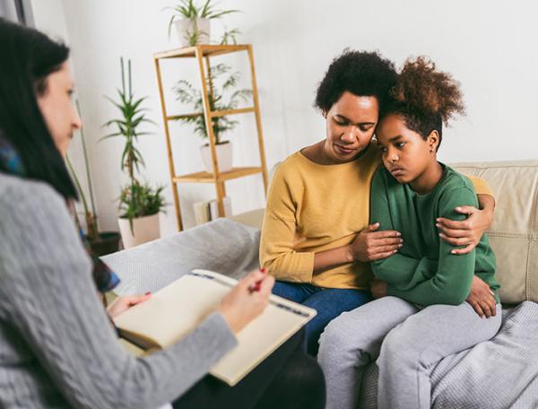 A mother and despondent looking child sit on a counselors sofa