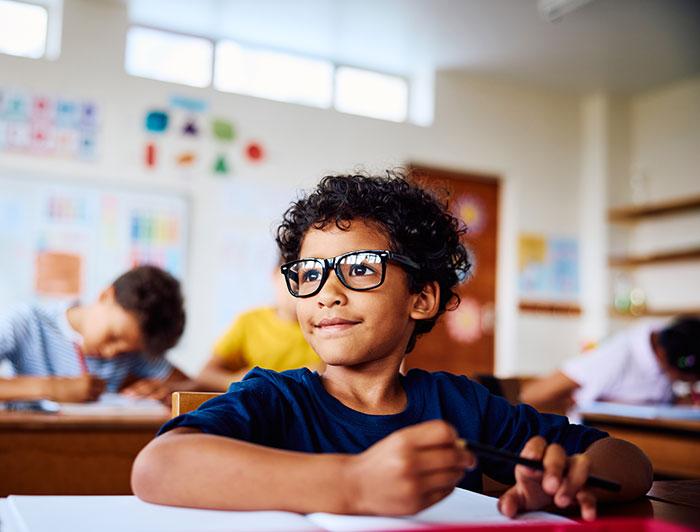 A boy wearing glasses sits at a school desk in a classroom